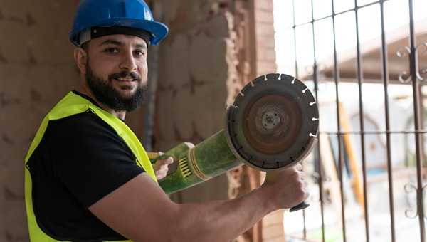 Construction worker using equipment while wearing a blue hard hat and yellow construction vest 